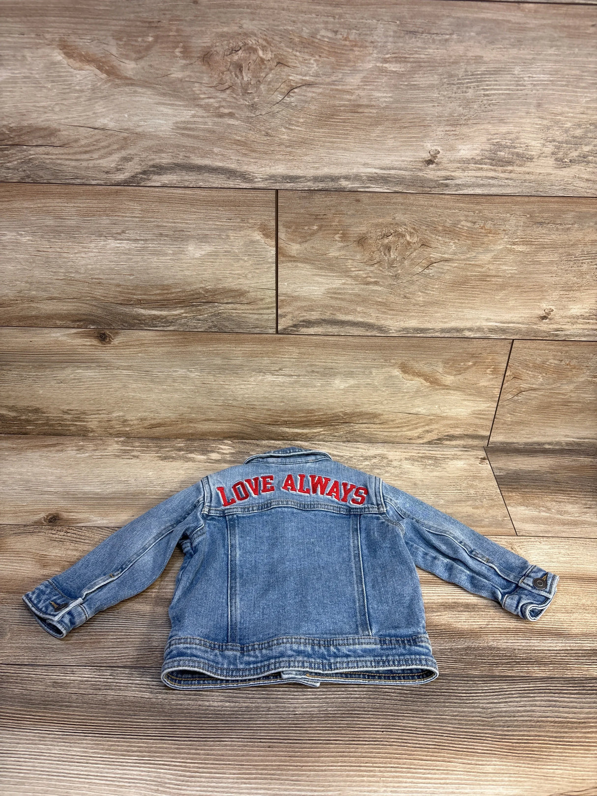 Kids denim jacket with 'Love Always' embroidered on back, laid flat on wood floor