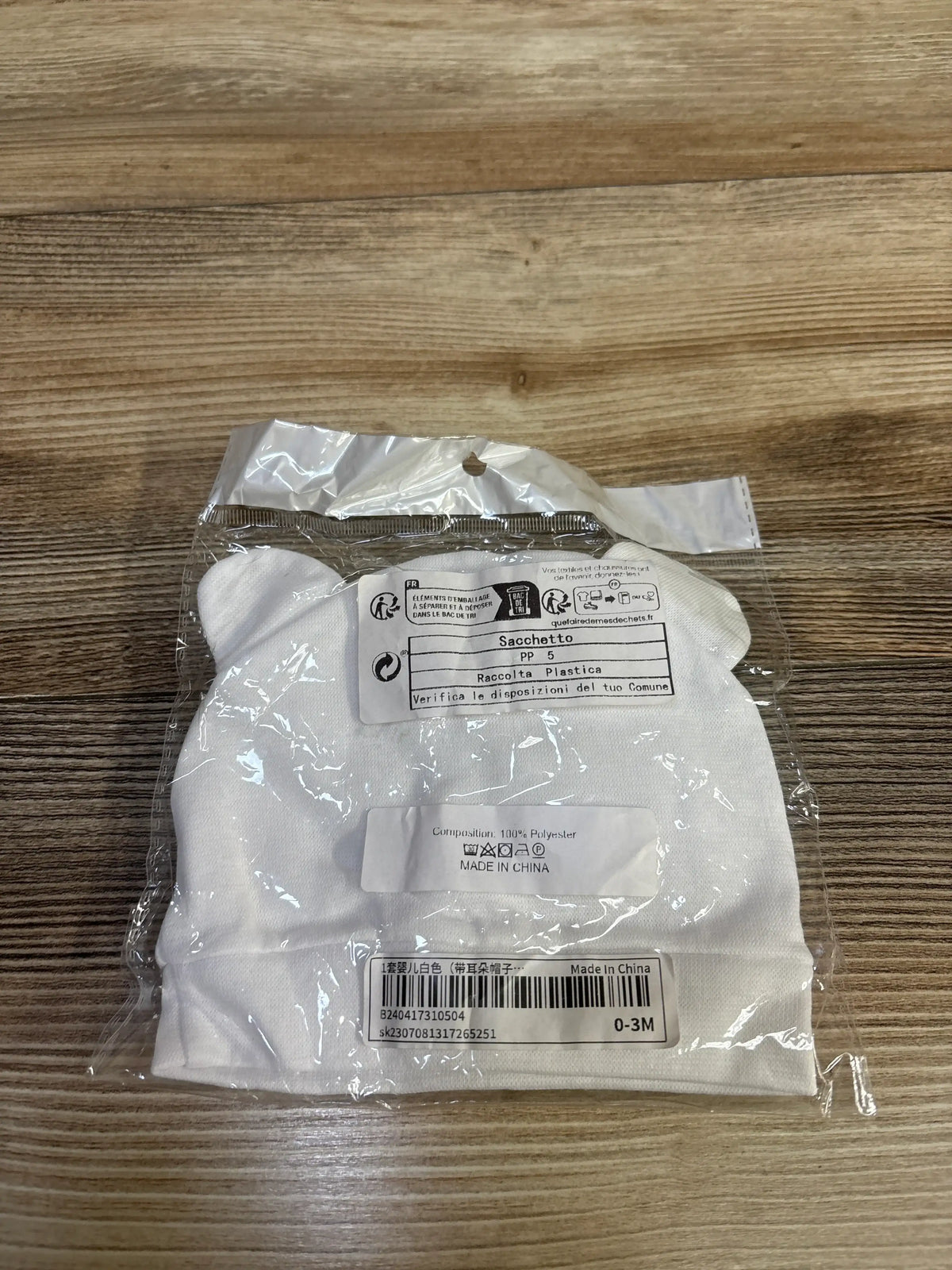 Top view of white Sacchetto newborn hat mittens and booties set in clear packaging on a wood surface