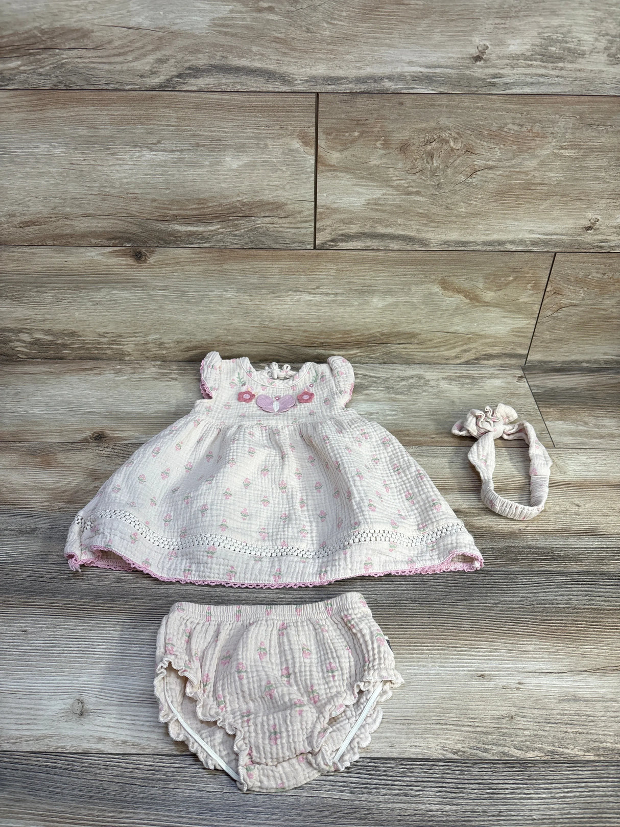 Flat lay of beige 3pc embroidered gauze dress set size 12 months showing smocked embroidered yoke, bloomers and headband on wood