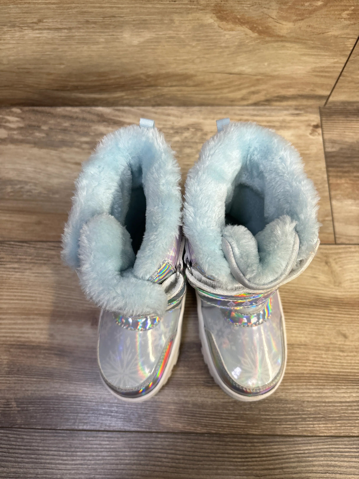 Top-down view of Disney Frozen toddler snow boots in holographic silver showcasing light blue faux-fur lining and front strap on wood floor