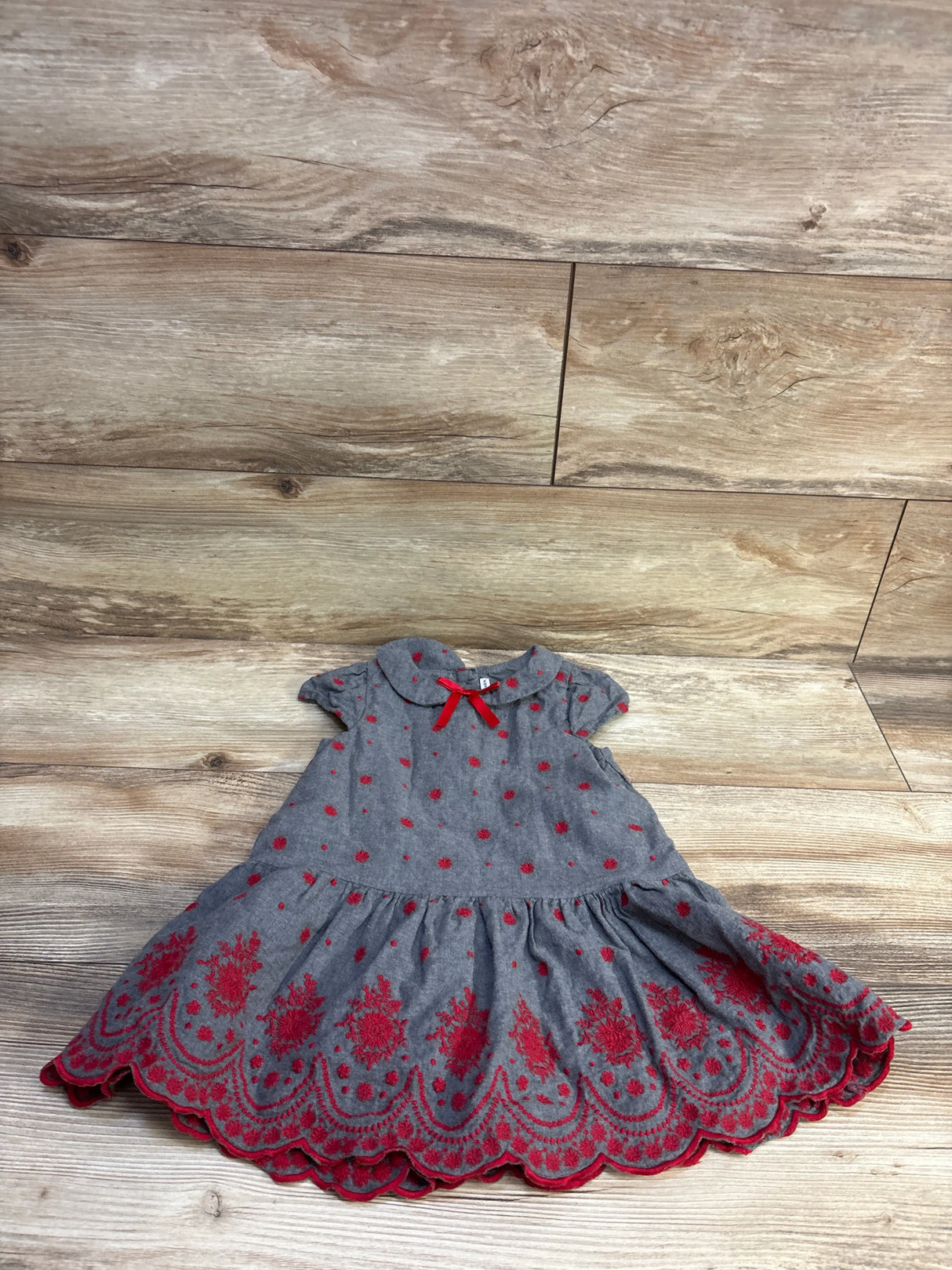 Front view of Janie & Jack The Festive Floral Embroidered Dress with red embroidery, scalloped hem, and bow on wood background
