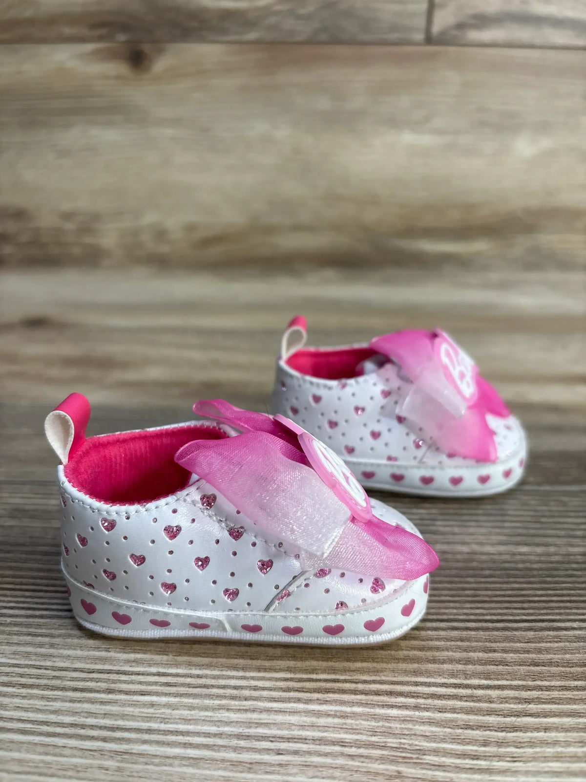 Angled front view of Barbie soft-sole baby sneakers size 1C with white and pink glitter hearts and pink organza bows on light wood