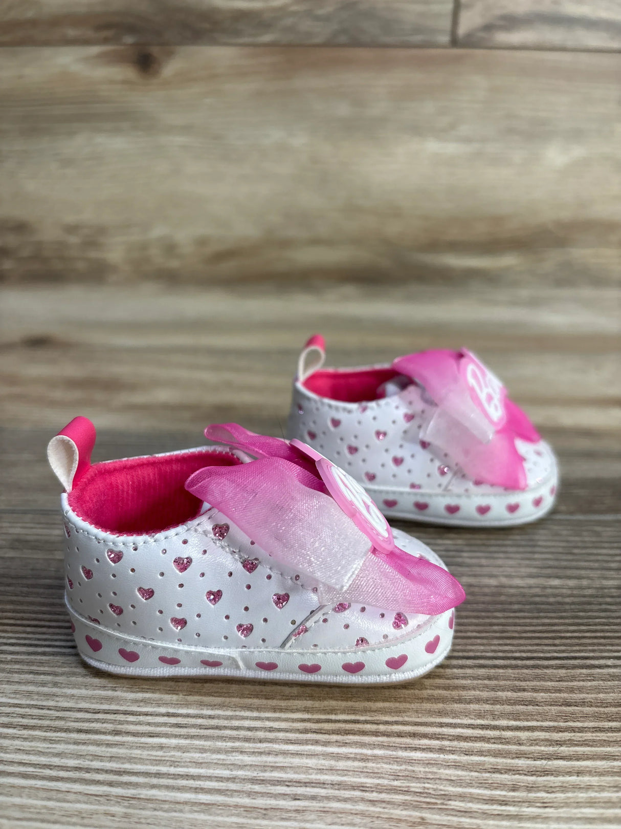 Angled front view of Barbie soft-sole baby sneakers size 1C with white and pink glitter hearts and pink organza bows on light wood