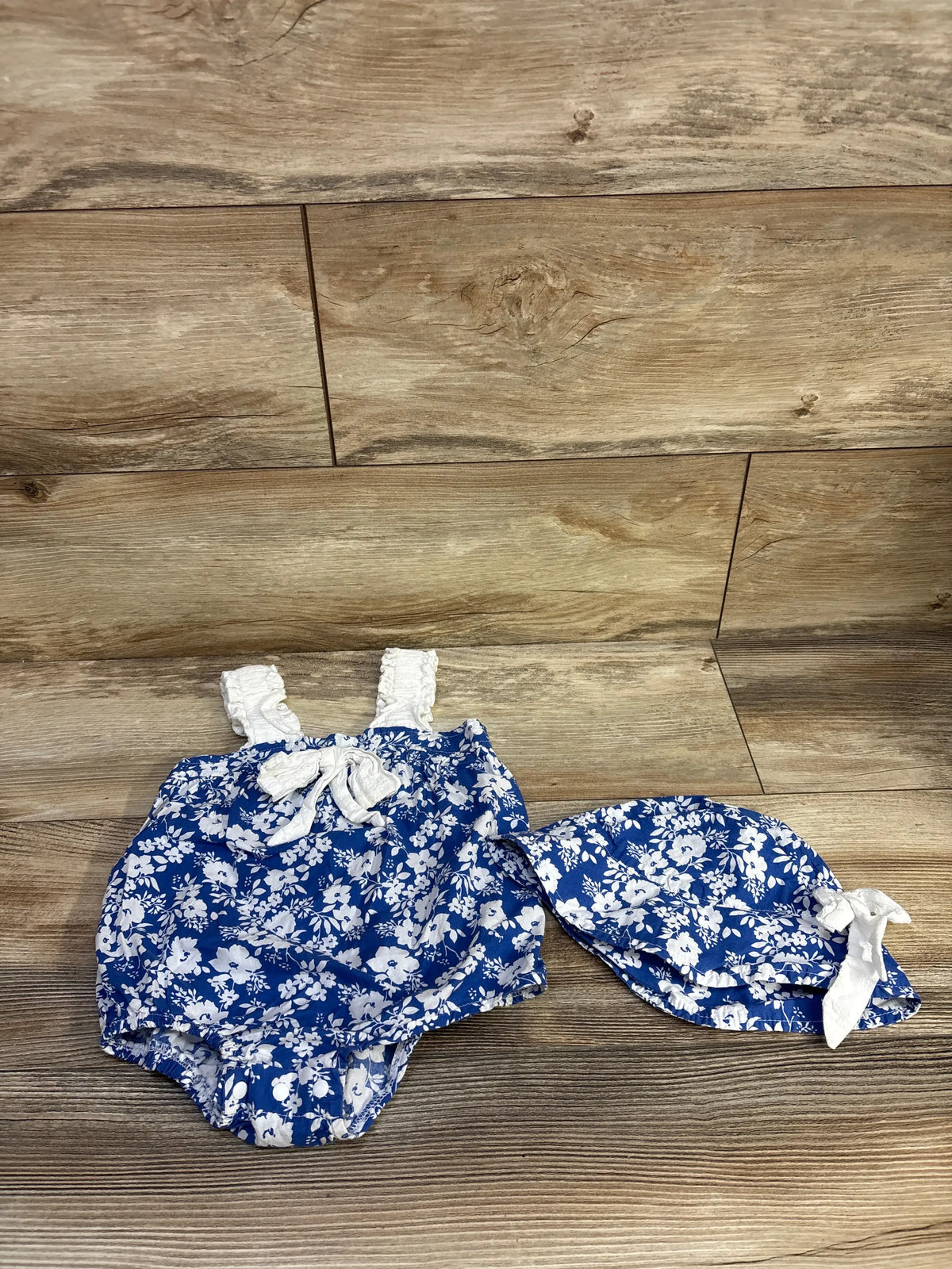 Top view of 2-piece blue floral bubble romper and hat set showcasing white straps and bow on a wood floor