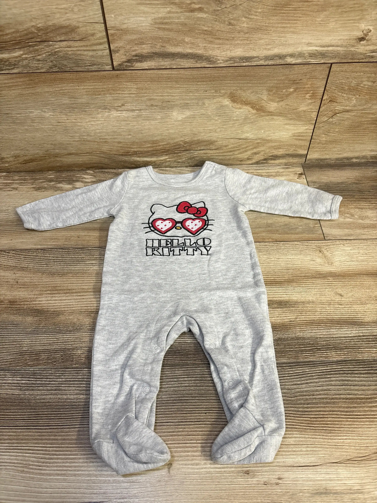 Front view of grey Hello Kitty footed coverall (6m) with heart-sunglasses Kitty print and snap leg closures on wood floor