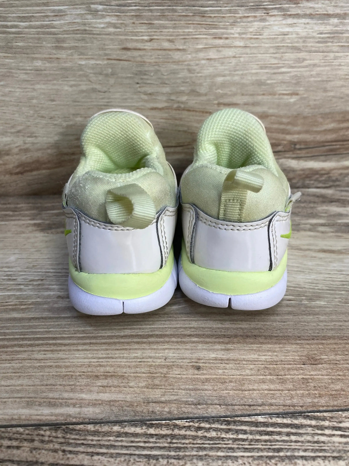 Rear view of toddler Nike Dynamo Free sneakers showing white patent heel and pale green lining with wood background