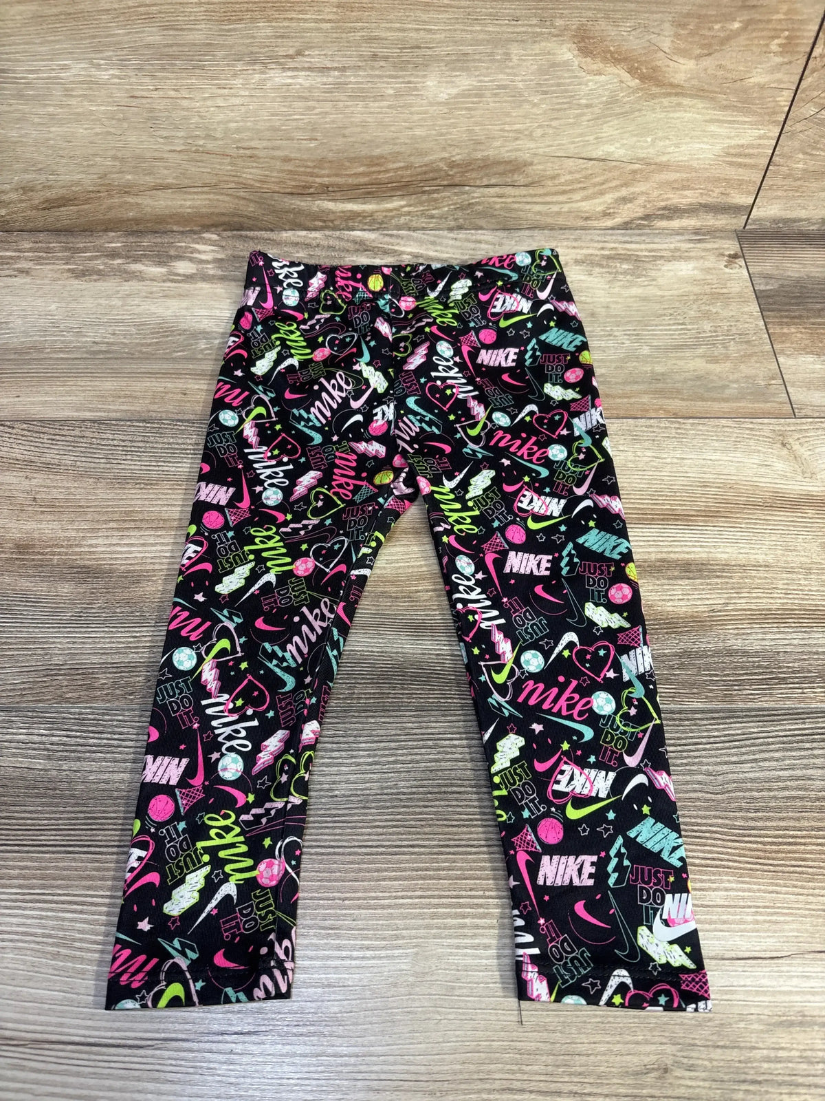 Flat lay of toddler Nike leggings, 24 months, showing colorful all-over Nike logos and graphics on wood floor