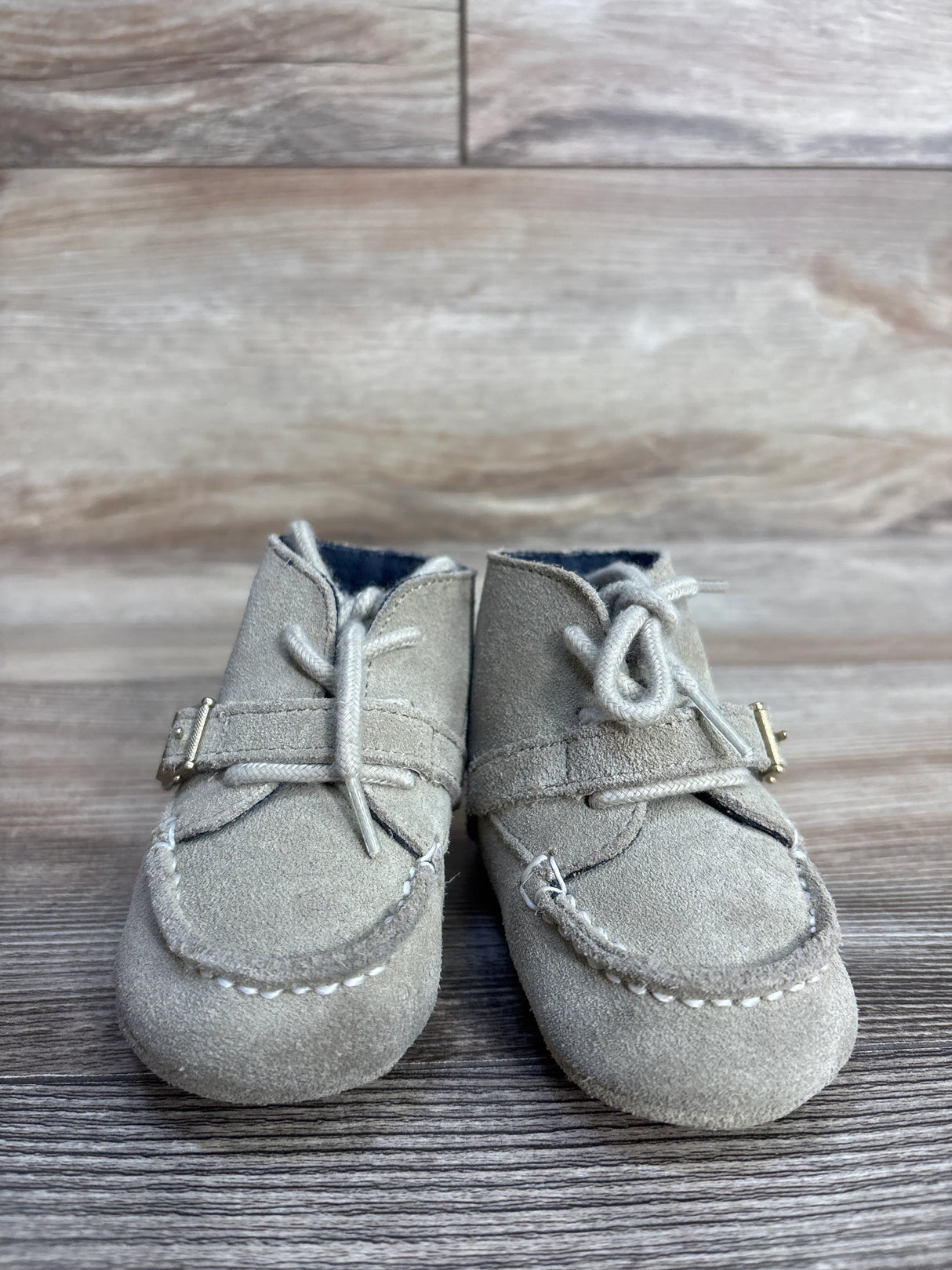 Pair of beige suede baby moccasin shoes with laces and buckle detail on wooden surface