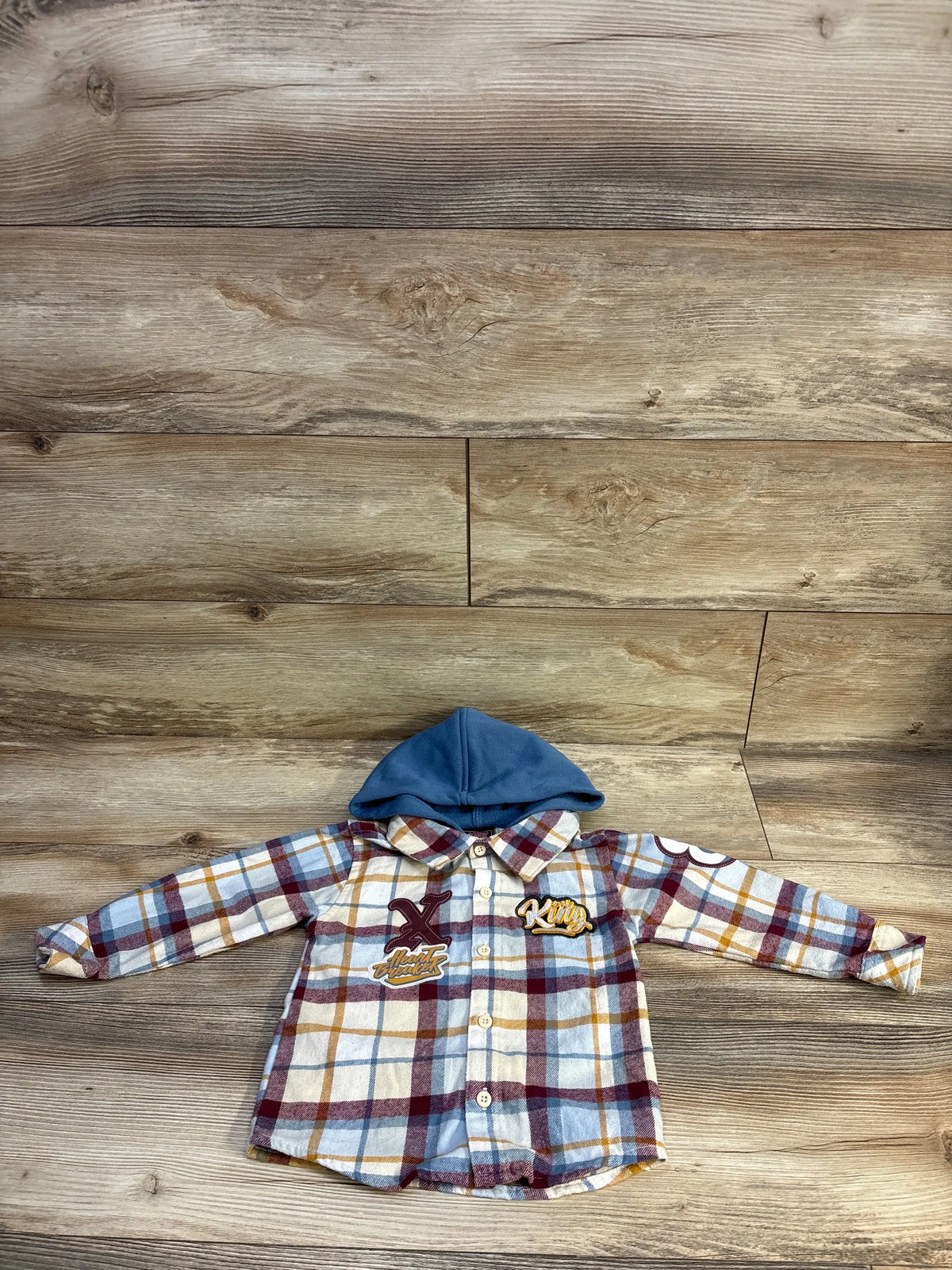 Front view of Mini Fresh hooded flannel in blue with plaid pattern, button front, hood, and graphic patches on wood background