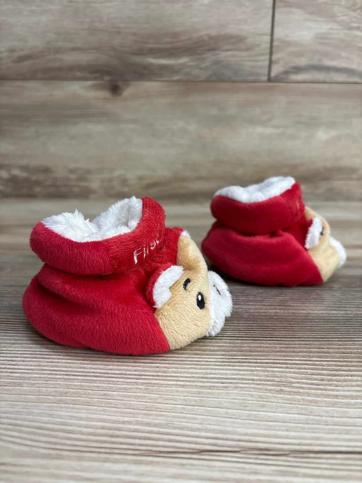 Red Santa-themed baby booties with plush white trim and cute face design on wood floor