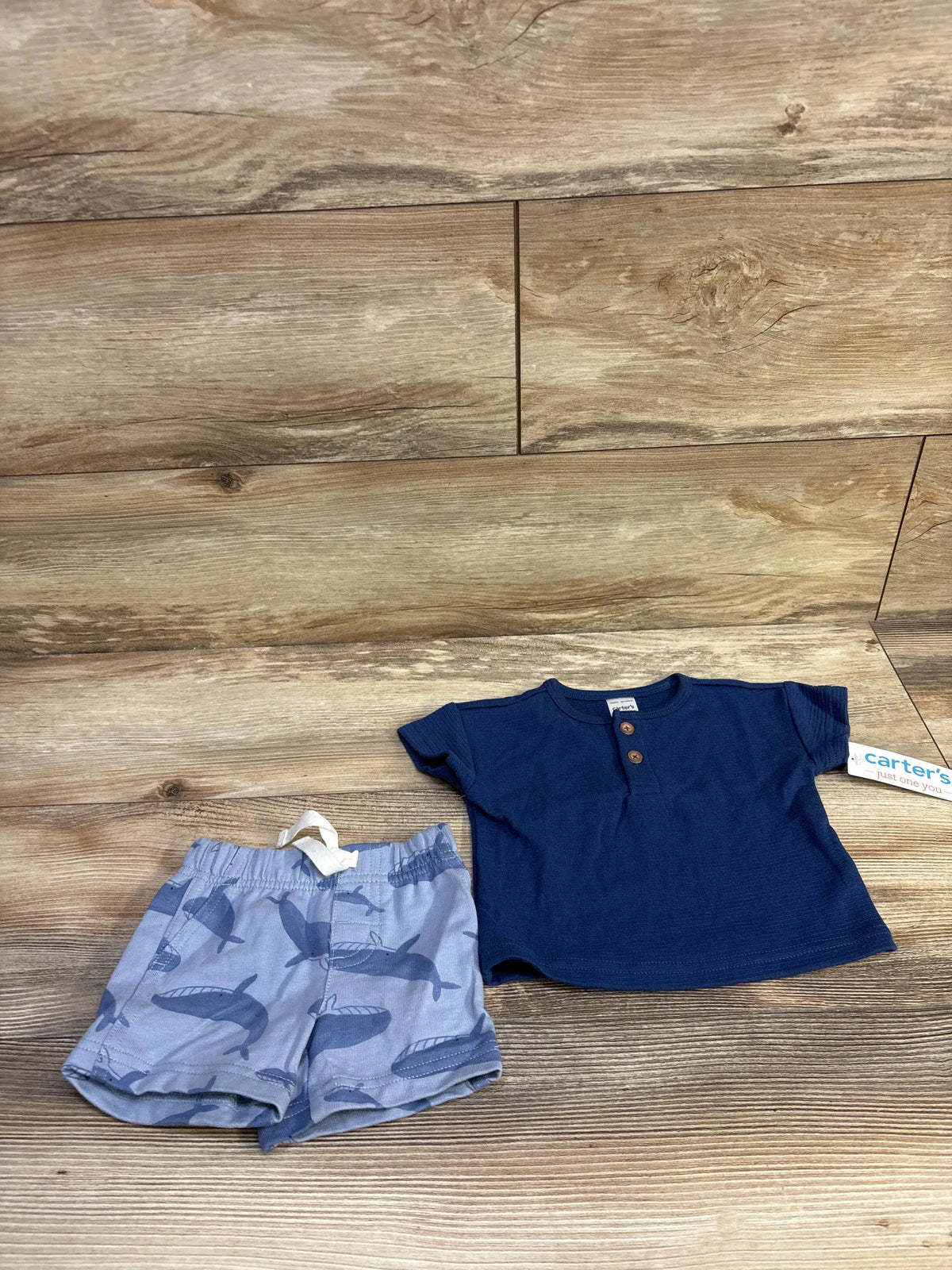 Flat lay of navy textured short-sleeve baby top with wooden buttons and light blue whale-print shorts, 3 months, on wood background