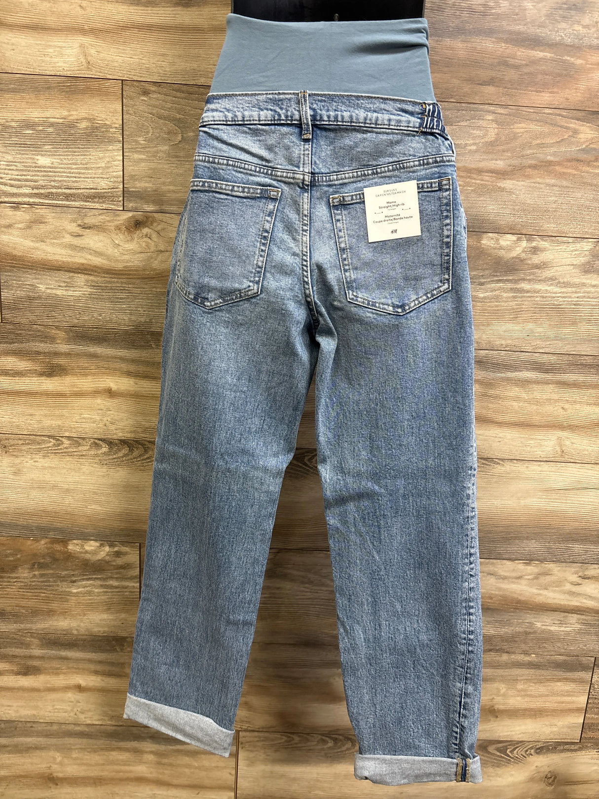 Rear view of H&M Mama maternity straight high-rib jeans showing high rib waistband, back pockets, and cuffed hems on a wooden floor background