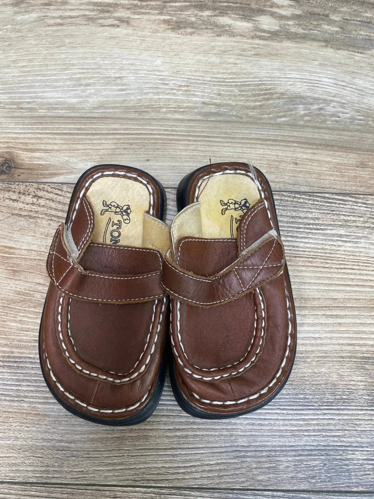 Overhead view of toddler Tom Dog brown leather clogs showcasing white contrast stitching and Velcro strap on wood floor