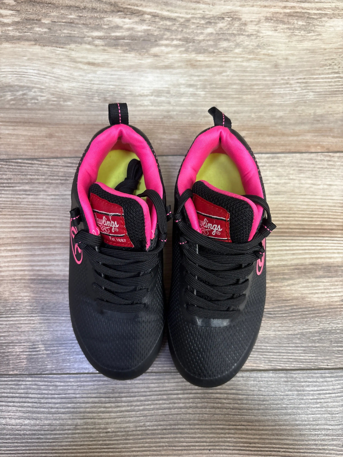 Top view of Rawlings girls softball cleats showcasing black uppers with pink lining and accents on a wood background