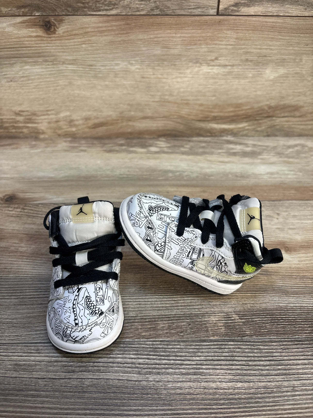 Front view of Jordan toddler high-top sneakers showcasing white uppers with black sketch graphic print and black laces on wood plank floor