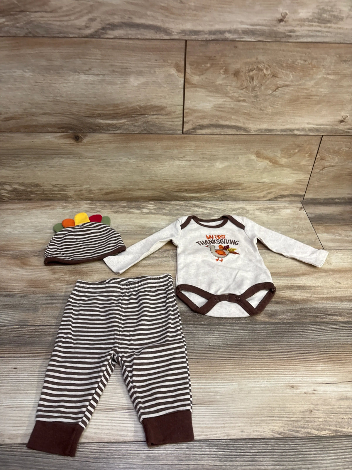 Top view of Baby Essentials My First Thanksgiving 3pc set with striped pants, hat, and turkey bodysuit on wood-look flooring