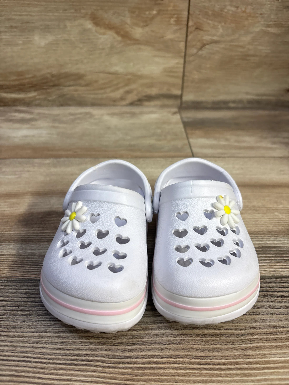 Front view of white Koala Kids clogs showing heart-shaped cutouts, daisy charms, and pink-striped soles on wood background