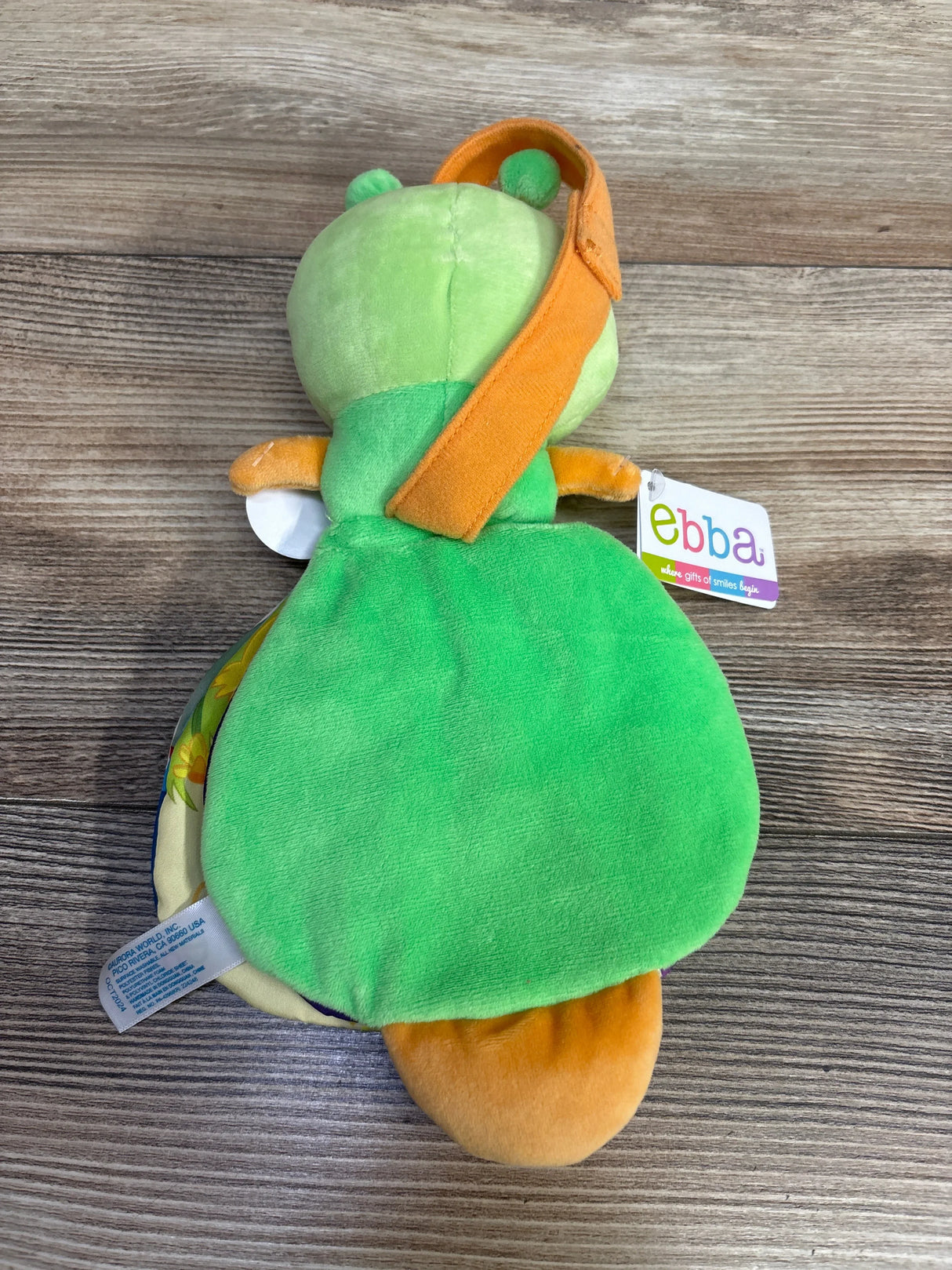 Rear view of ebba Baby Talk Story Pals Color is Fun plush book showing green frog back, orange hanging strap and ebba tag on wood background