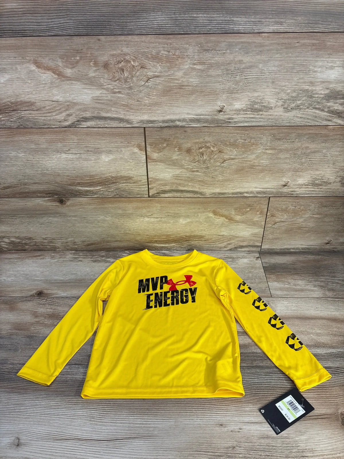 Flat lay front of yellow Under Armour long-sleeve shirt showing MVP ENERGY chest print, black sleeve graphics, tag on wood