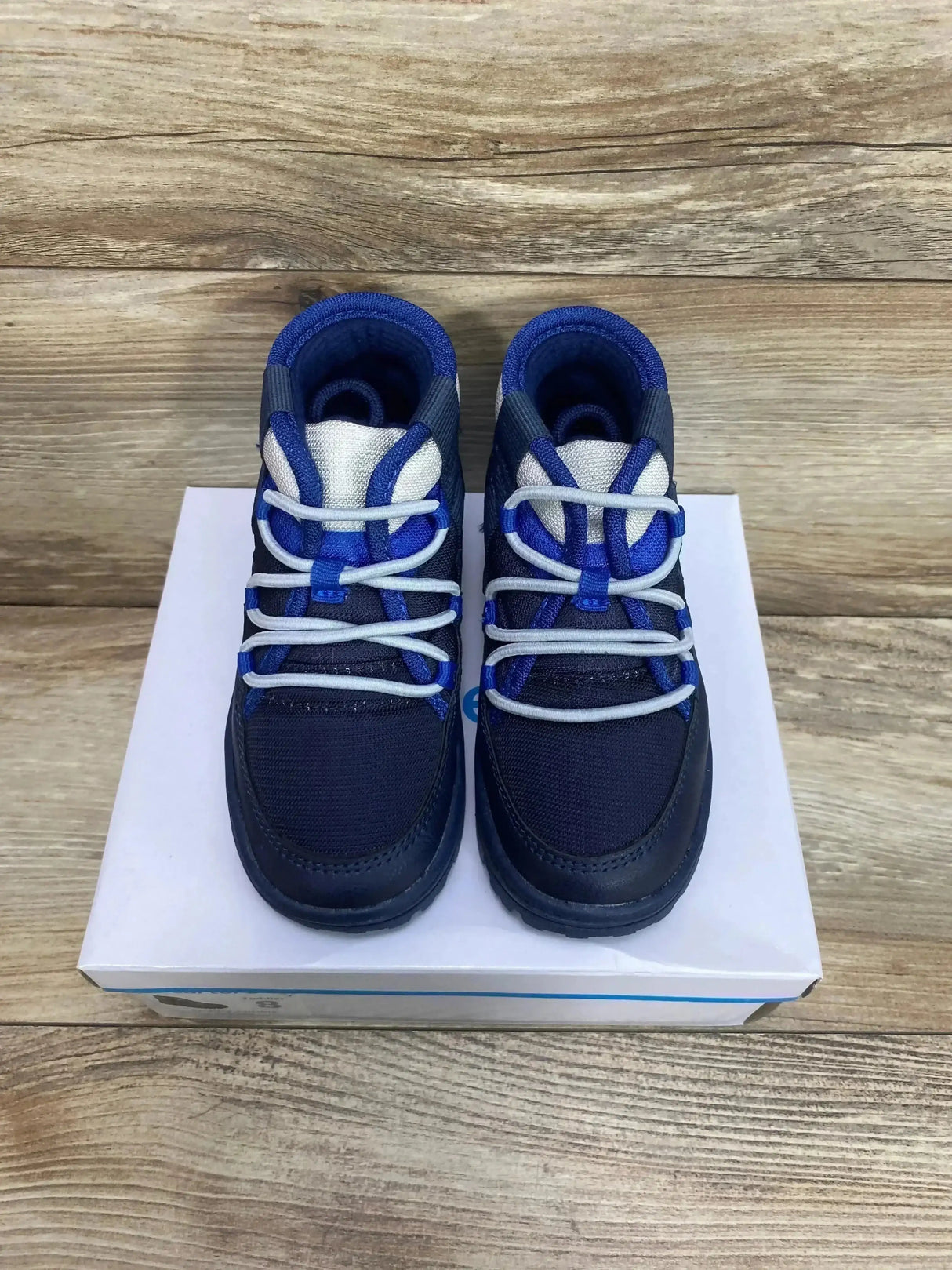 Top down view of Carter’s toddler Fallon boots showcasing white elastic laces and royal blue trim on a white shoebox over wood background
