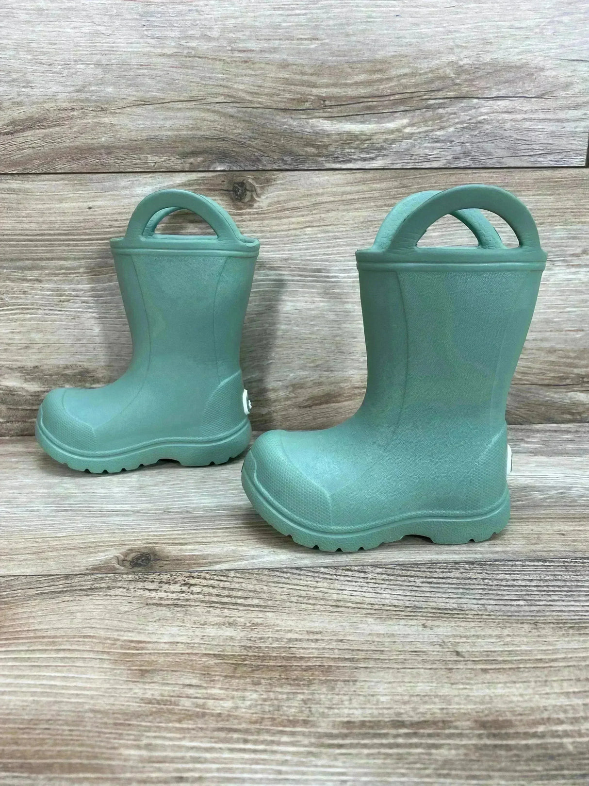 Three quarter view of toddler green BEARPAW EVA rain boots showing pull on handles and lugged soles on wood plank background