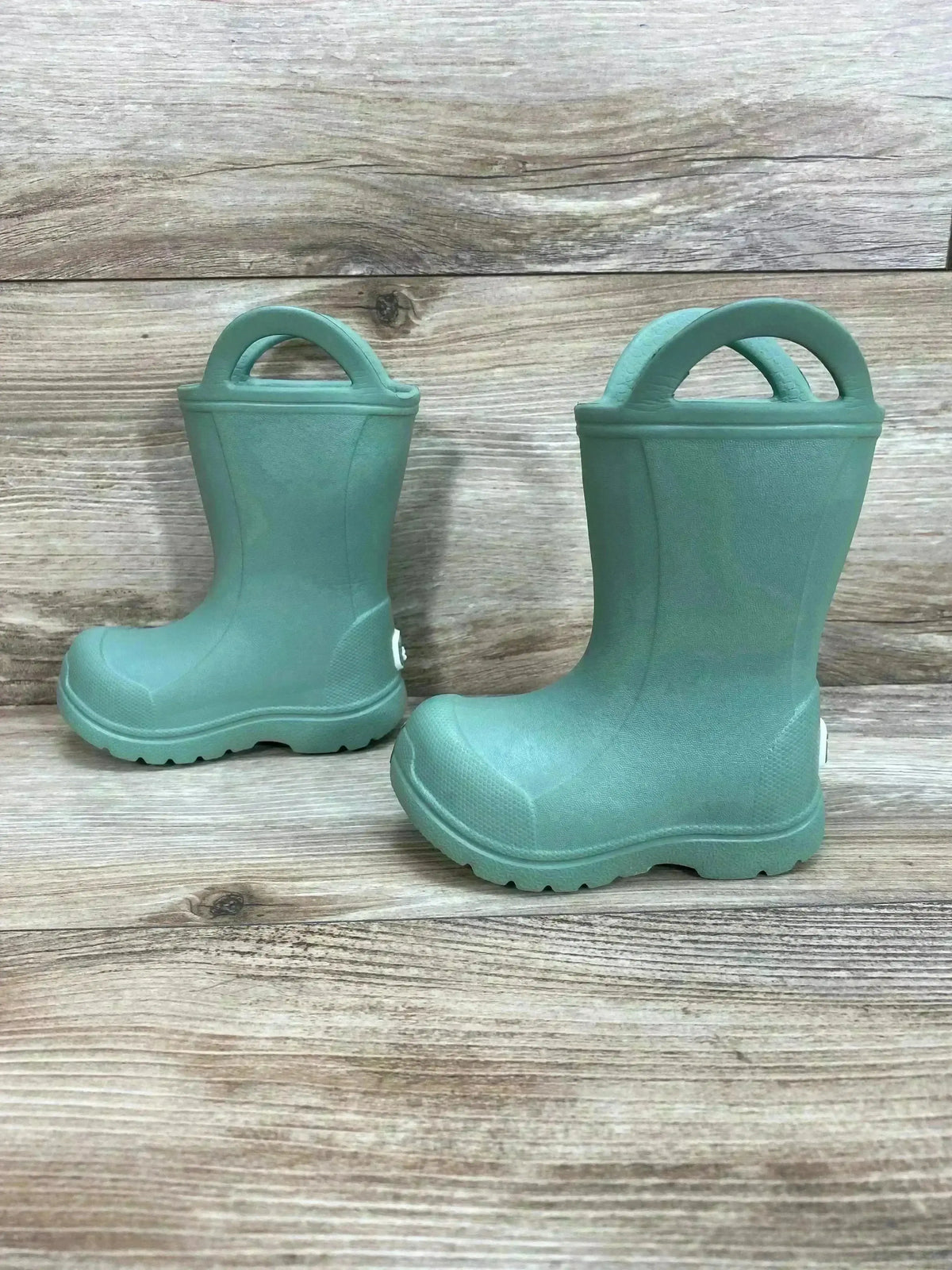 Three quarter view of toddler green BEARPAW EVA rain boots showing pull on handles and lugged soles on wood plank background