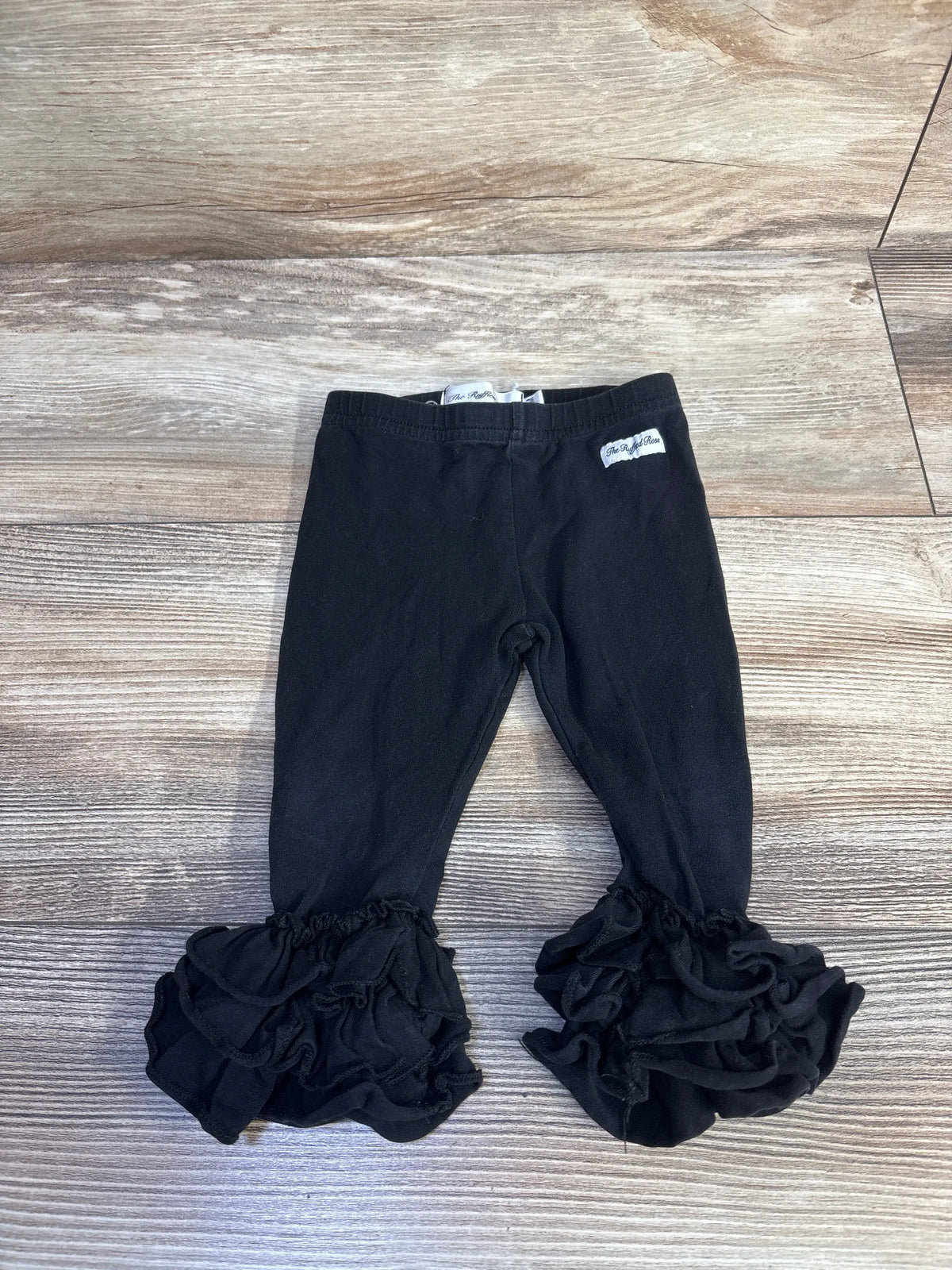 Flat lay of The Ruffled Rose black leggings in size 12 months showcasing layered ruffled cuffs on a wood-grain floor