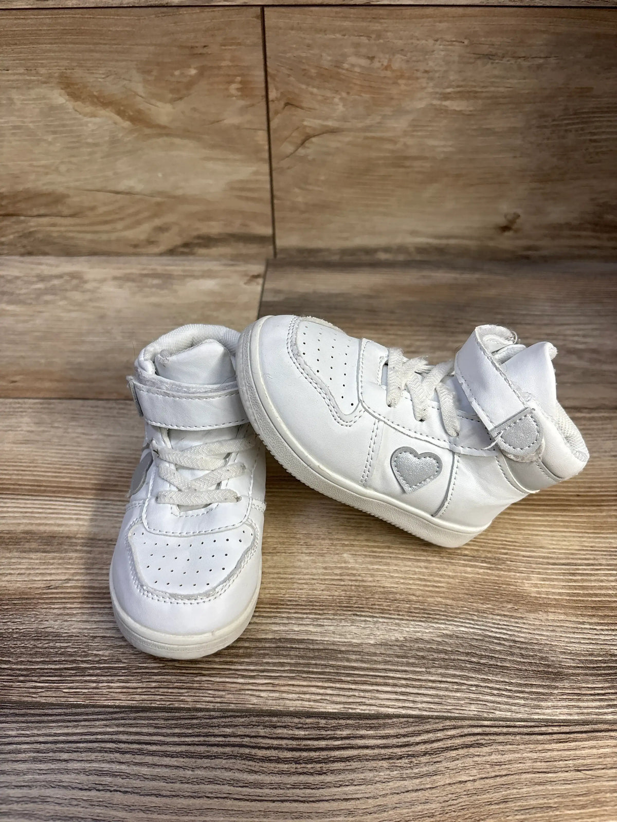 Front-left view of white First Steps high-top baby sneakers showing heart patch, Velcro strap, and perforated toe on wood background