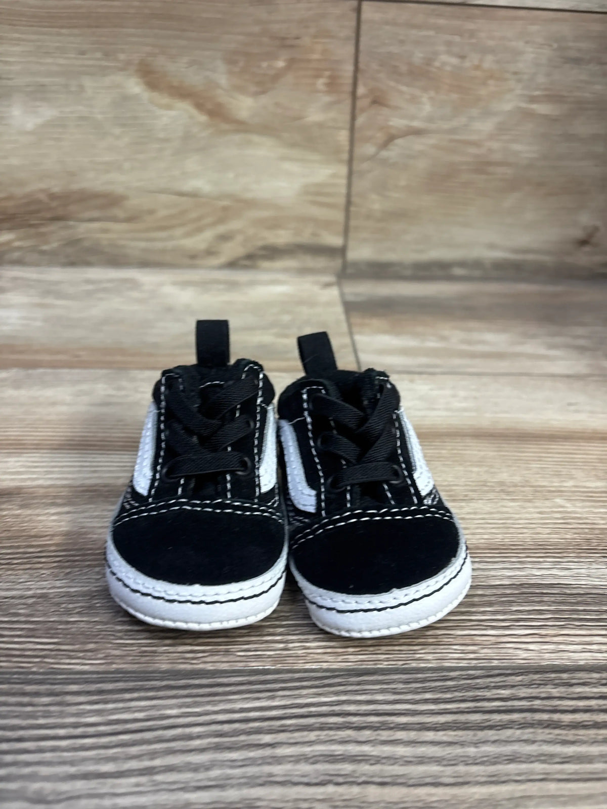 Front view of Vans Old Skool crib shoes in black showcasing white side stripes, laces, and white soles on a wood-look floor