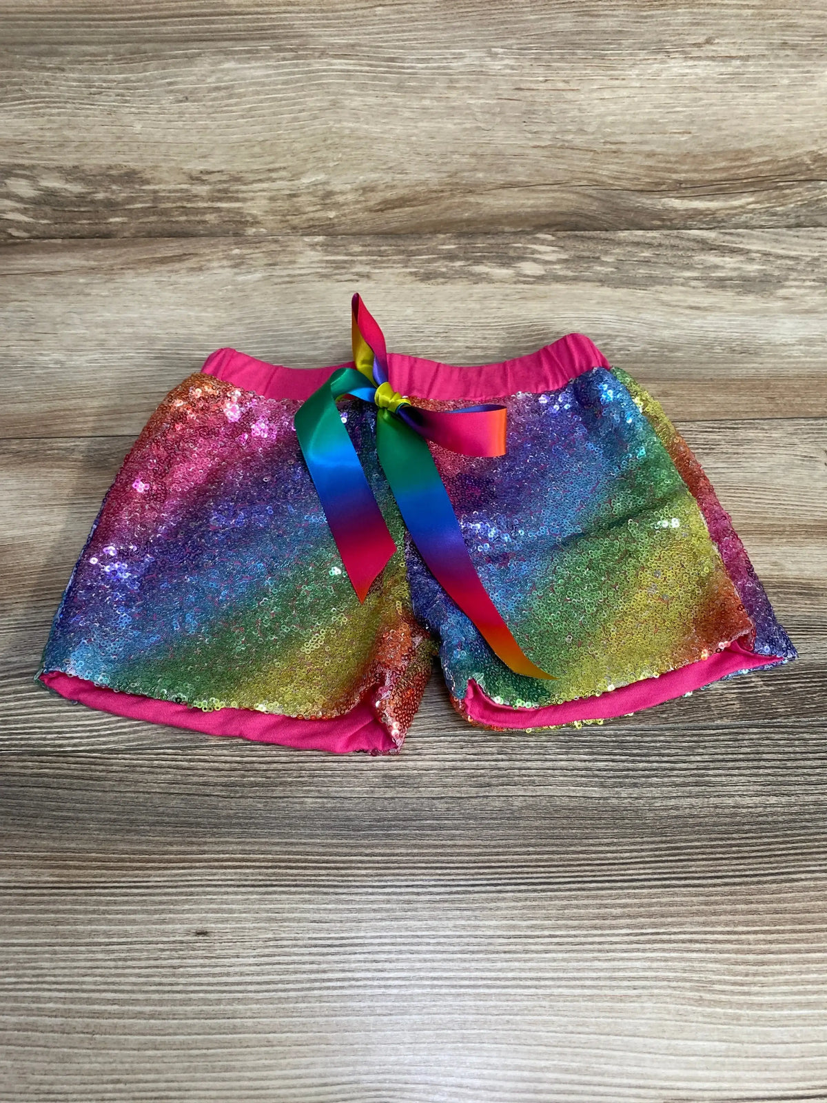 Front view of toddler rainbow sequin shorts showcasing multicolor sequins, rainbow ribbon bow at pink elastic waistband on wood background