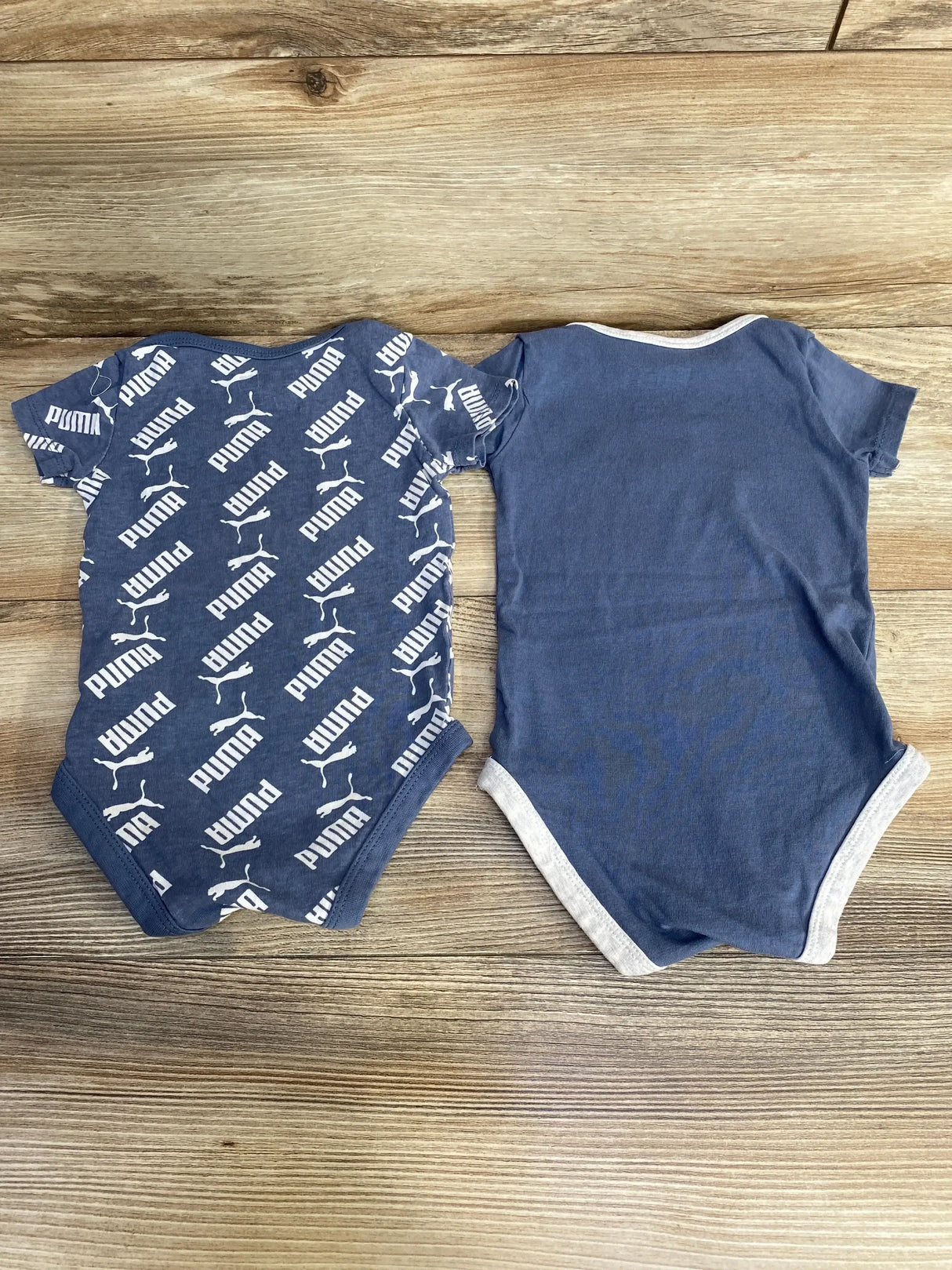 Top-down view of two blue Puma baby bodysuits showcasing one with all-over white Puma logo print and one solid blue with gray trim on a light wood background