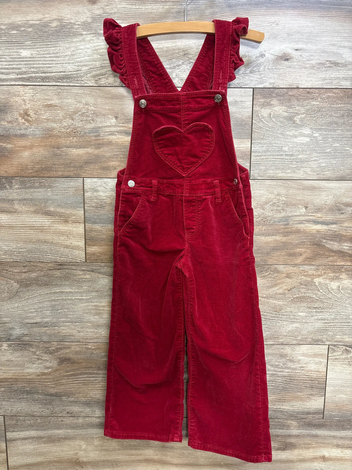 Front view of Baby Gap red velvet heart overalls showcasing ruffle straps, heart bib pocket, and slant pockets on a wood floor