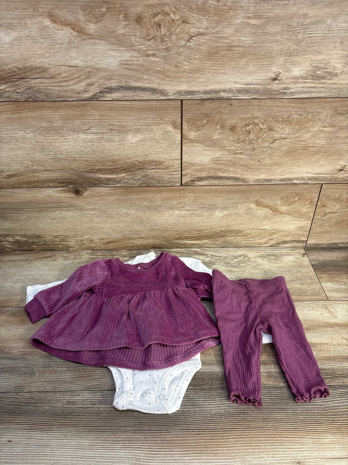 Flat lay of a three-piece baby set showing a mauve velvet ruffle peplum top, white snap bodysuit, and leggings on wood