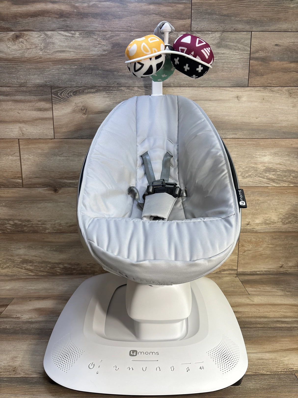 Front view of 4moms mamaRoo baby swing showing grey seat, five-point harness, toy mobile, and white base on wood floor