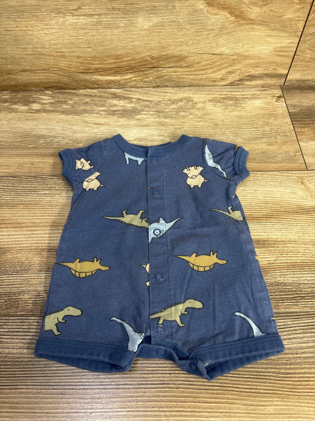 Front view of Carter's newborn blue shortie romper showcasing all-over dinosaur print and front snap closures on wood background