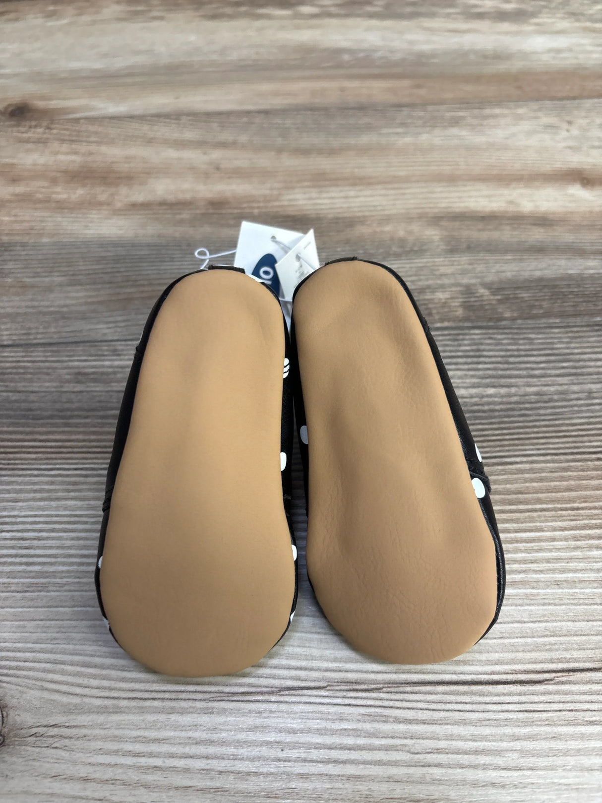 Pair of black baby shoes with white polka dots and soft tan soles on wooden surface
