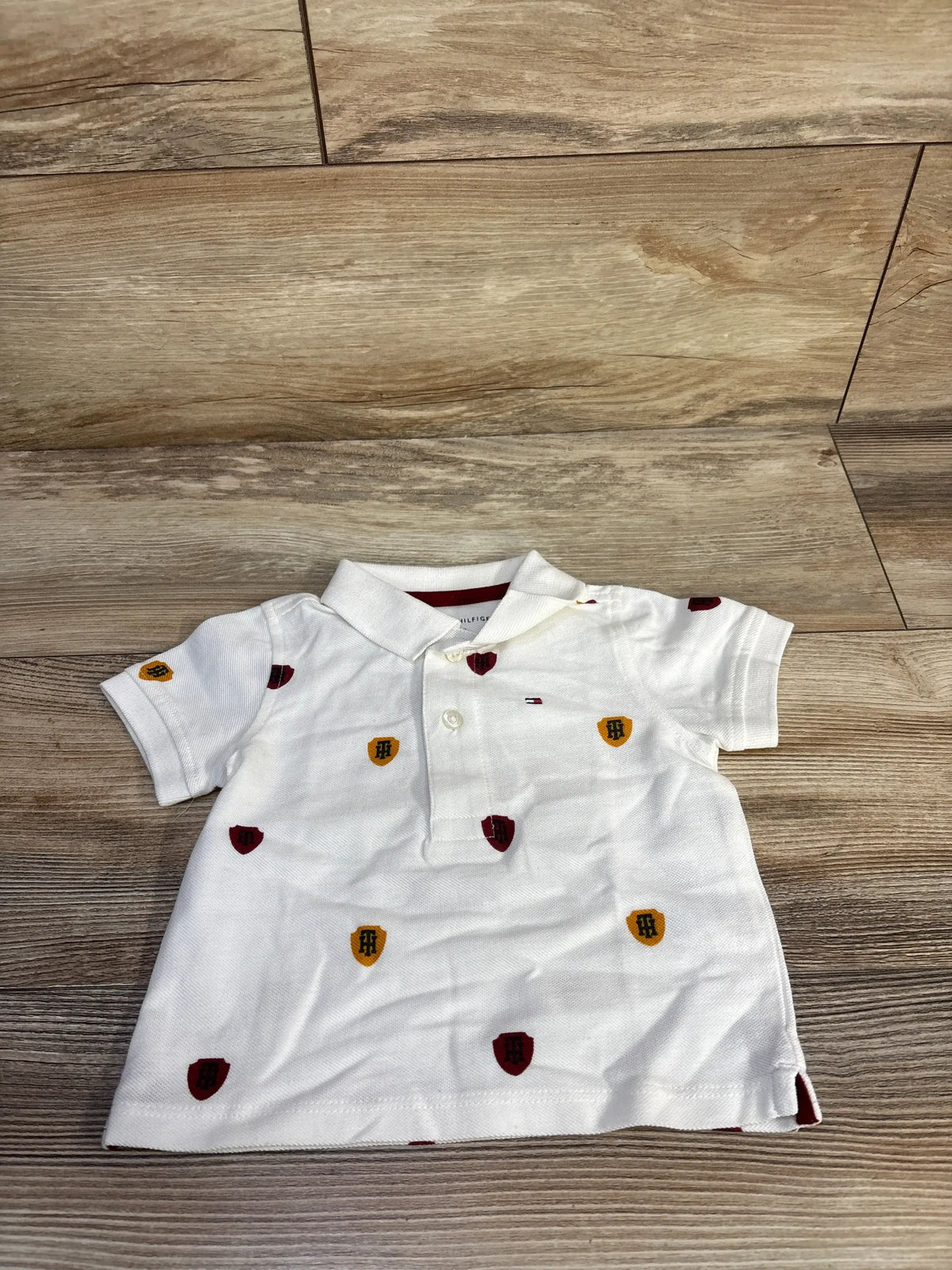 Front flat lay of white Tommy Hilfiger polo shirt with embroidered red and yellow shields and flag logo on light wood