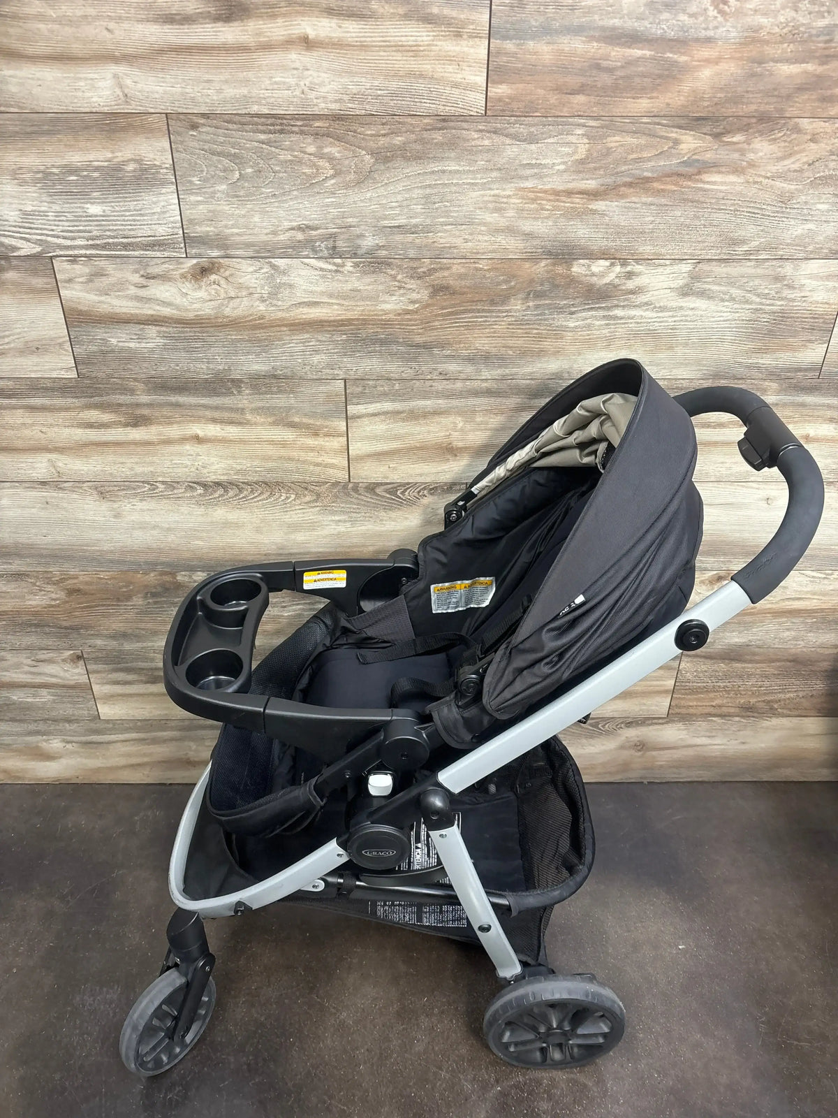 Angled view of Graco Modes Pramette Stroller showcasing black seat, canopy, cup holder, and storage basket on wood flooring