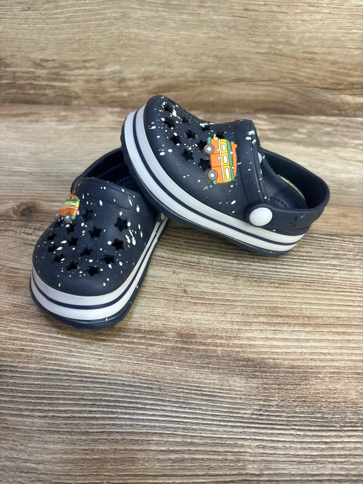Three-quarter view of Koala Kids blue star cut-out clogs showcasing star-shaped holes, white paint-splatter and striped sole on wooden backdrop