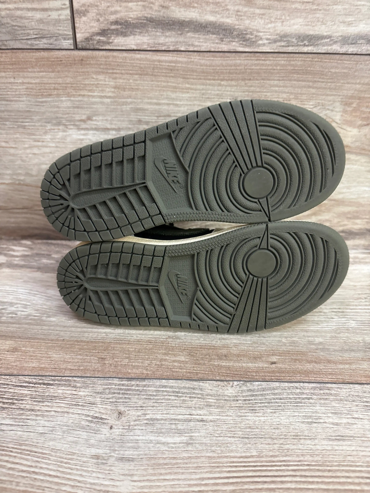 Bottom view of Travis Scott and Air Jordan 1 Retro Low OG SP PS Olive sneakers showcasing textured Nike Air outsoles on a wood-grain background