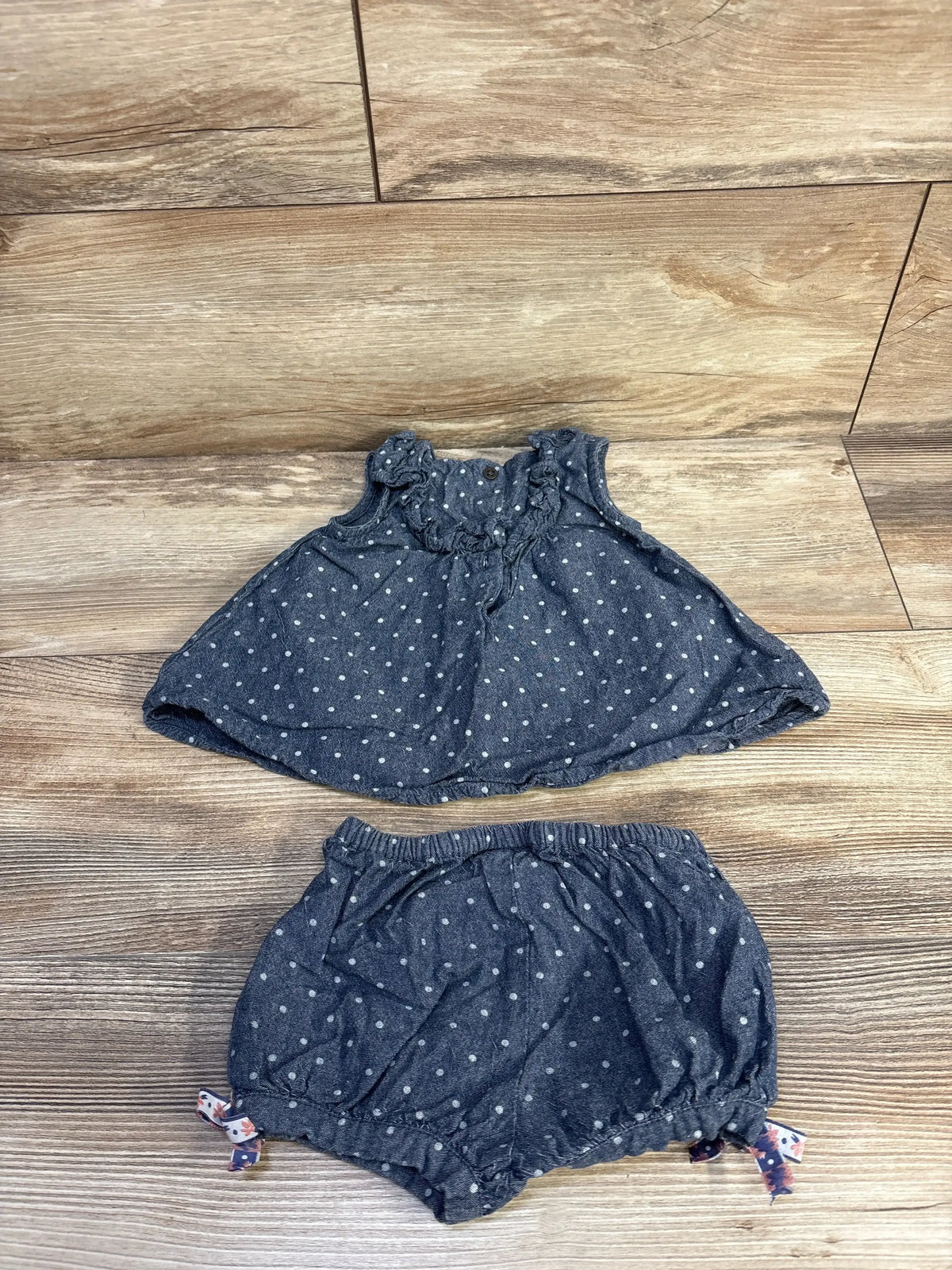 Flat lay of blue chambray baby top and bloomers showing white polka dots, ruffled neckline, elastic waist and tied leg bows on wood floor