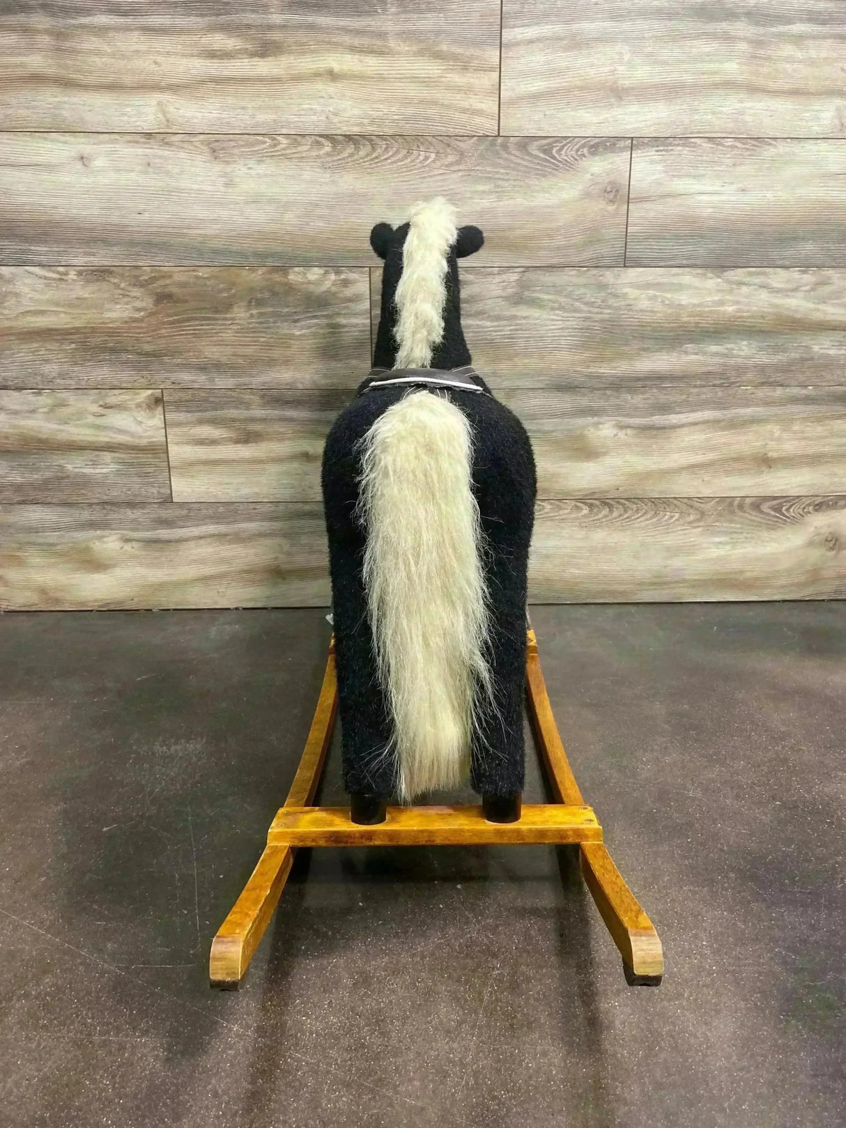 Rear view of vintage black plush rocking horse on wooden rocker, showing long cream mane and tail with wood plank backdrop