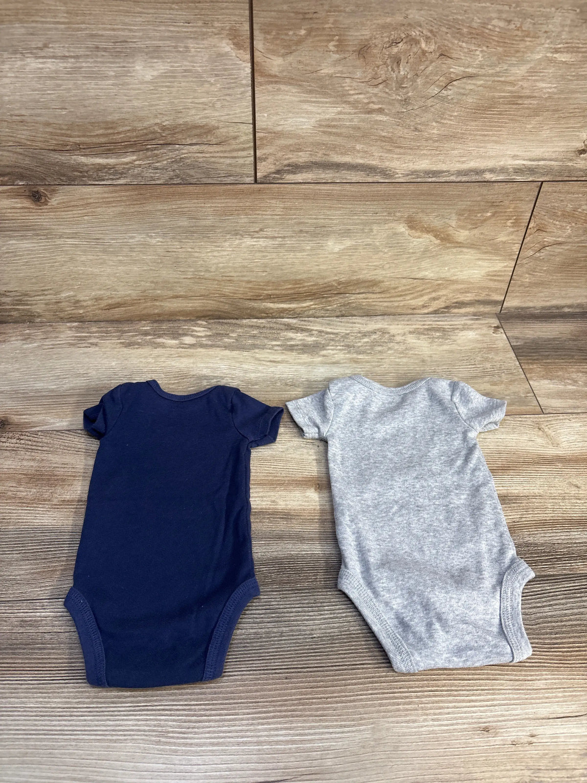 Front view of Just One You 2pk newborn bodysuits showcasing short sleeves and navy and grey colors on a wood surface