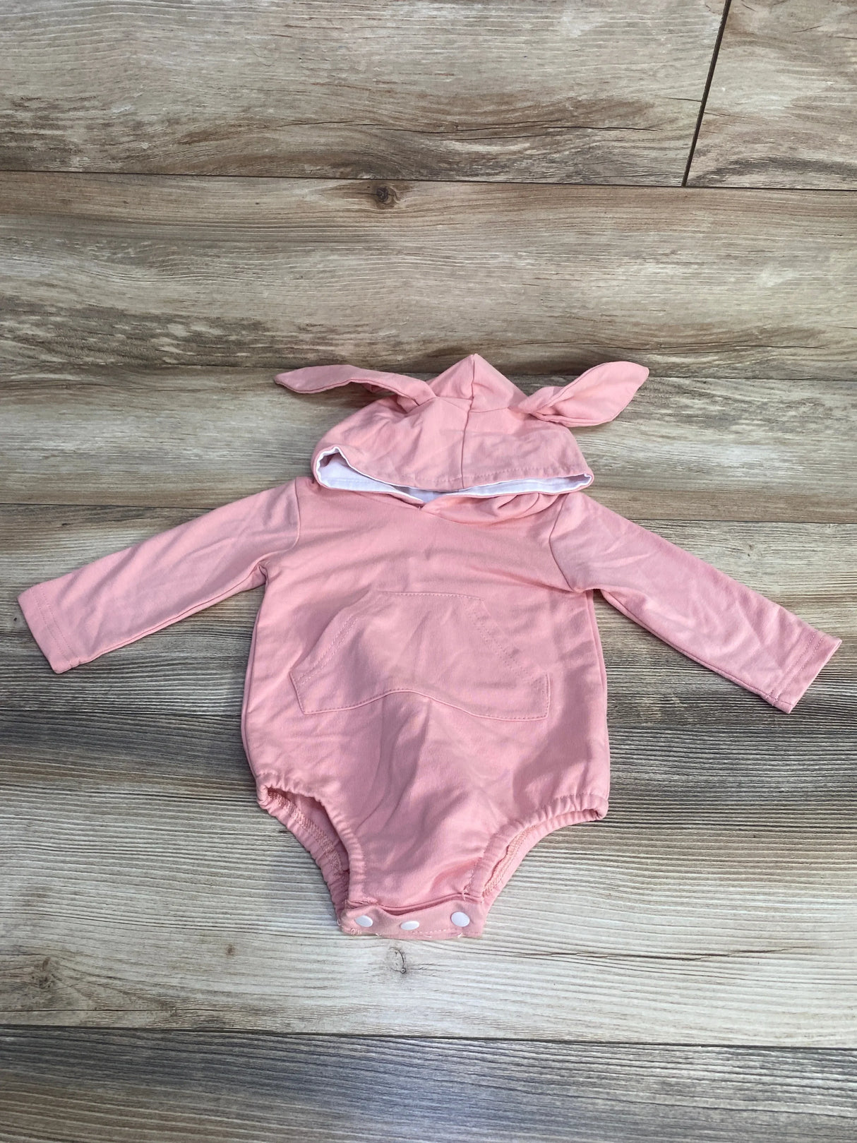 Flat lay of pink bunny hooded romper size 9–12 months showing bunny-ear hood, front pocket, long sleeves, and snap crotch on light wood floor