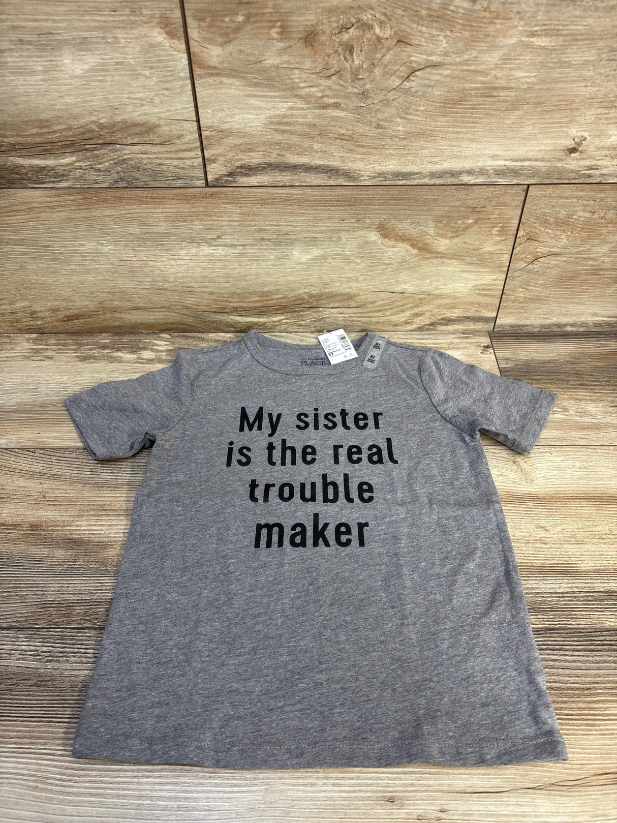 Gray kids t-shirt with 'My sister is the real trouble maker' text, size 5T, laid on wood floor