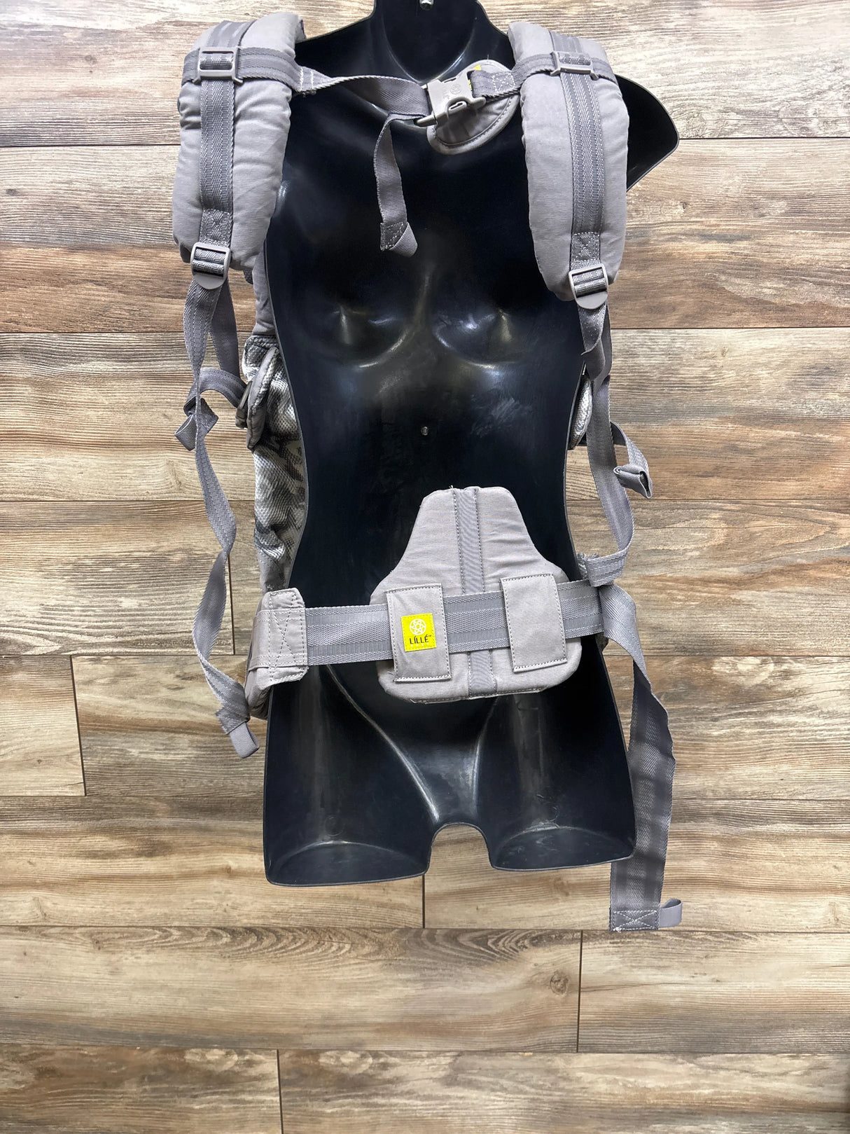 Gray LÍLLÉbaby baby carrier with padded straps on a mannequin, wood background