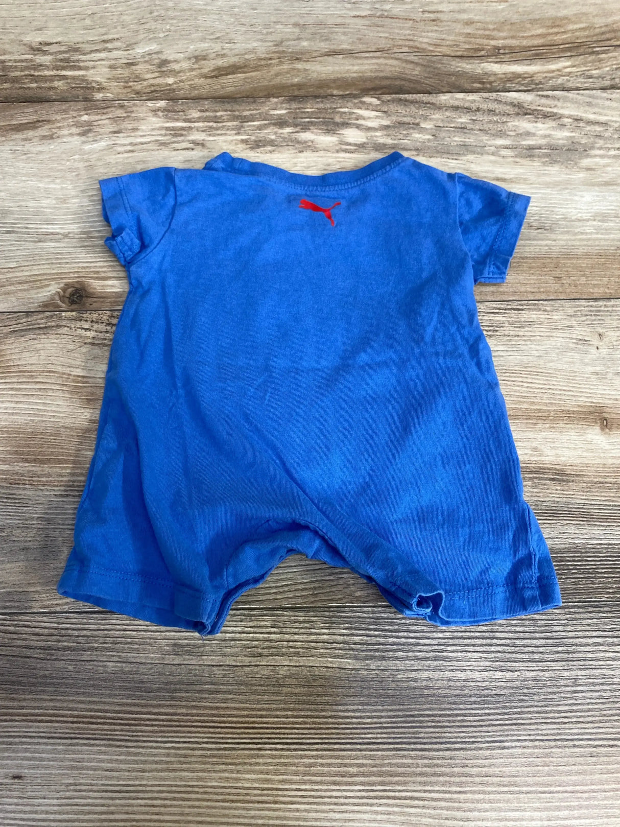 Flat lay of blue Puma shortie romper showing red Puma logo on upper back, short sleeves and snap leg openings on wooden floor