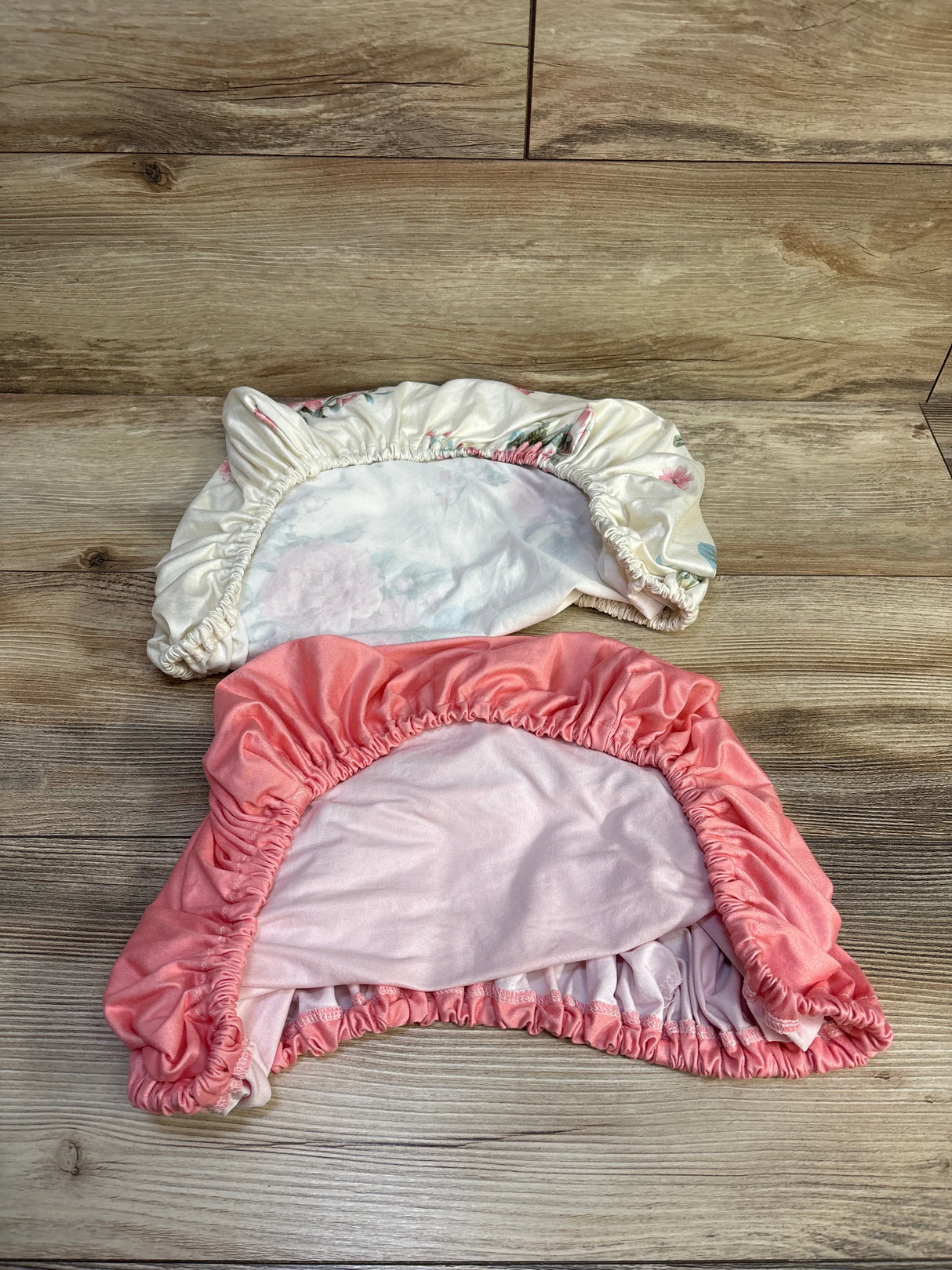 Flat lay of two fitted bassinet sheets, floral and pink, showing elastic edges, 23x36 in on wood background
