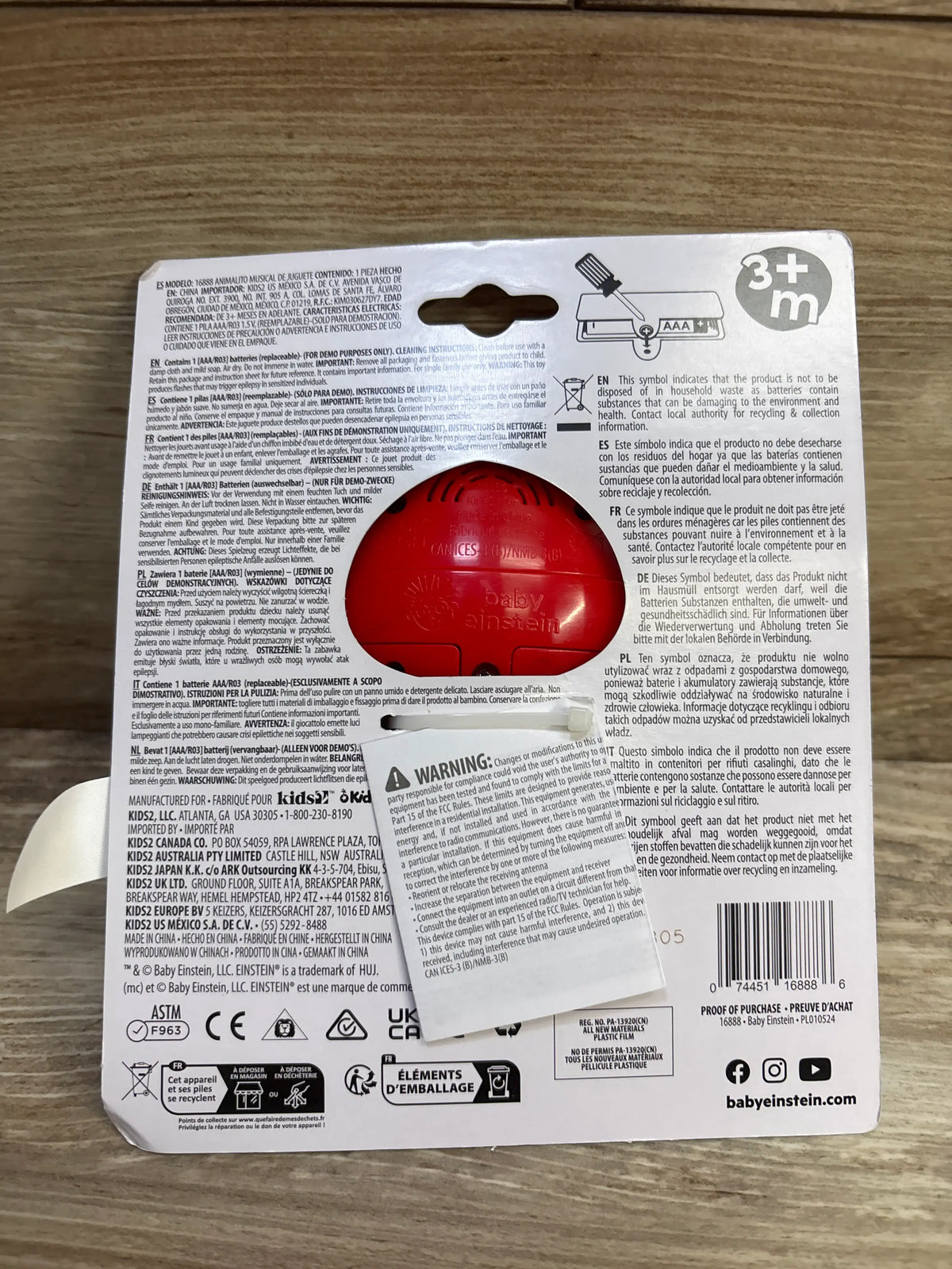 Back view of Baby Einstein Roxy’s Bright Flight Musical Toy showing the red toy unit inside retail packaging on a wood surface