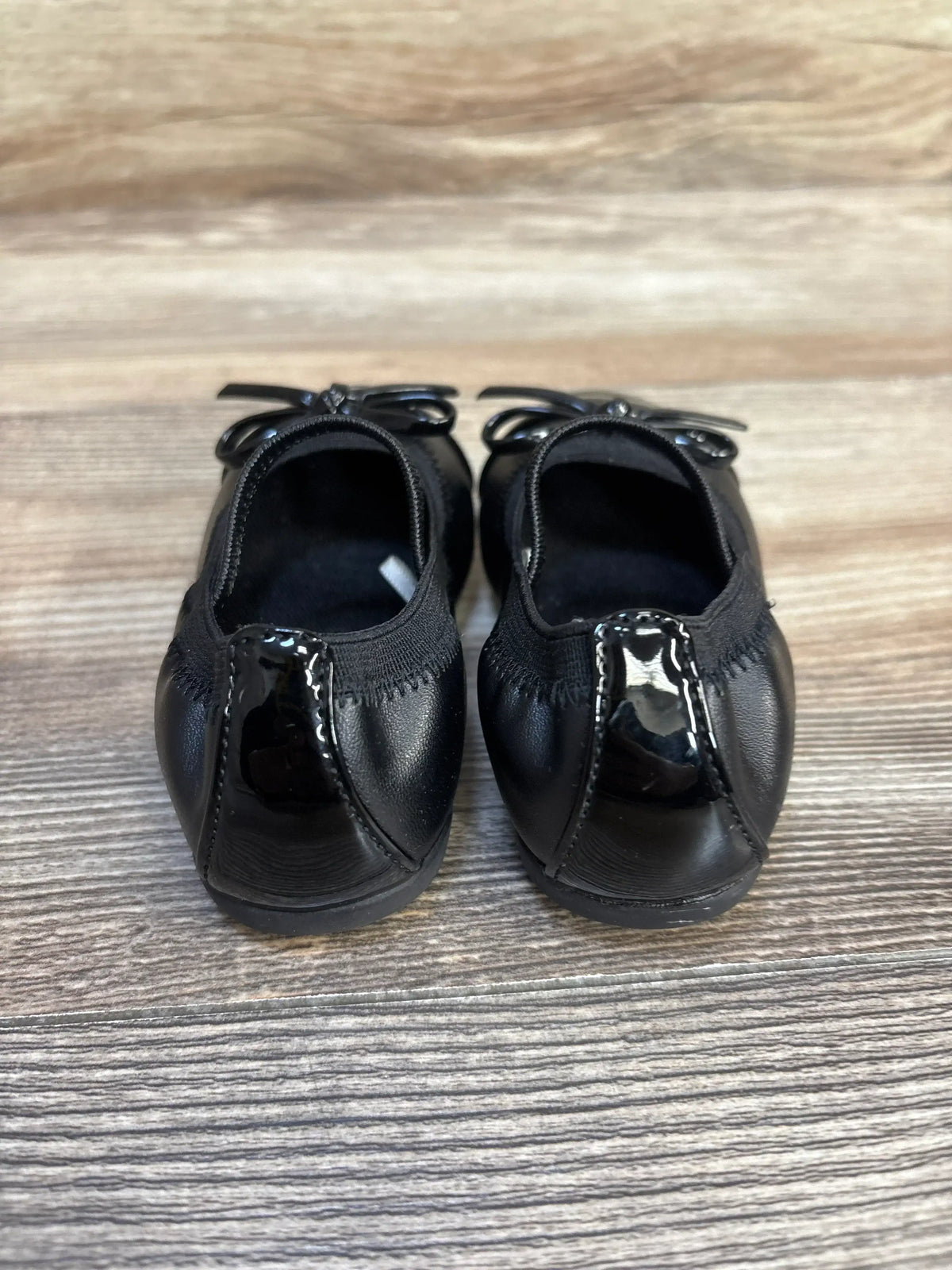 Back view of Children's Place black Mary Jane bow flats showcasing patent leather heels and elastic backs on wood plank floor